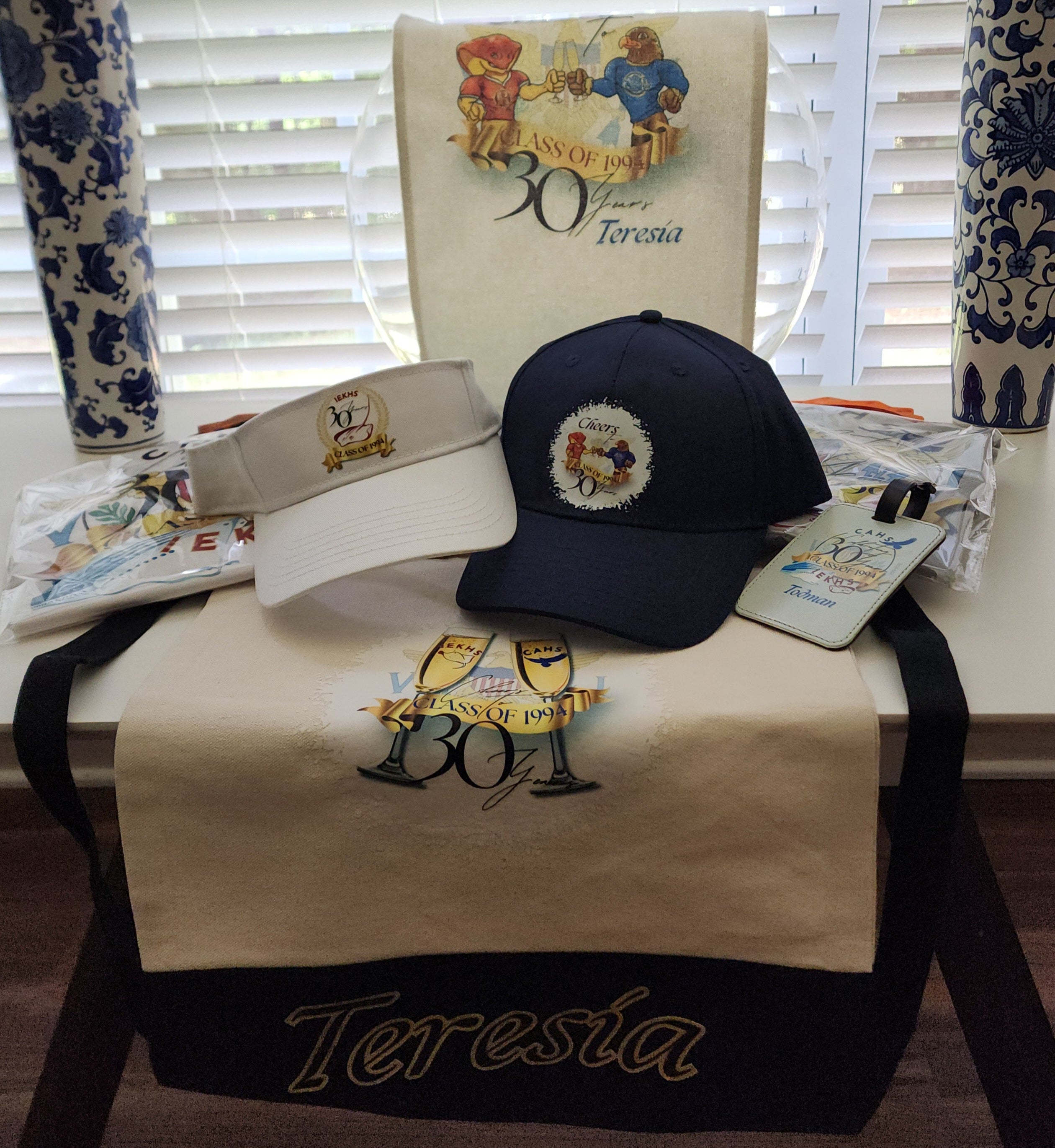 Table with branded items for a 30th Reunion including a cap, visor, tees, and towels on a table with decorative vases in the background.
